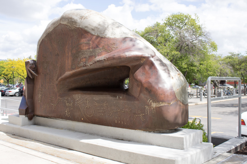 A weathered and stainless steel sculpture that depicts a Native American clinging to a rock formation that features petroglyphs. 