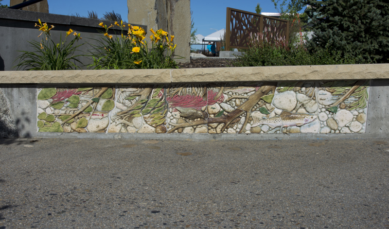 A flower bed retaining wall bench with a base decorated with painted low bas-relief ceramic sculptures depicting several fish swimming in a river. 