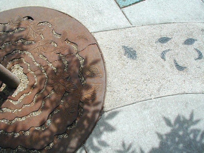 A series of patinaed bronze leaves inlayed in pea gravel aggregate cement that curves within the regular cement of a city sidewalk.  The aggregate cement ends at the end of a rusty iron tree grate that has wavy lines resembling water ripples. In between the ripple are low-relief sculpted leaves that match the bronze leaves in the cement. 