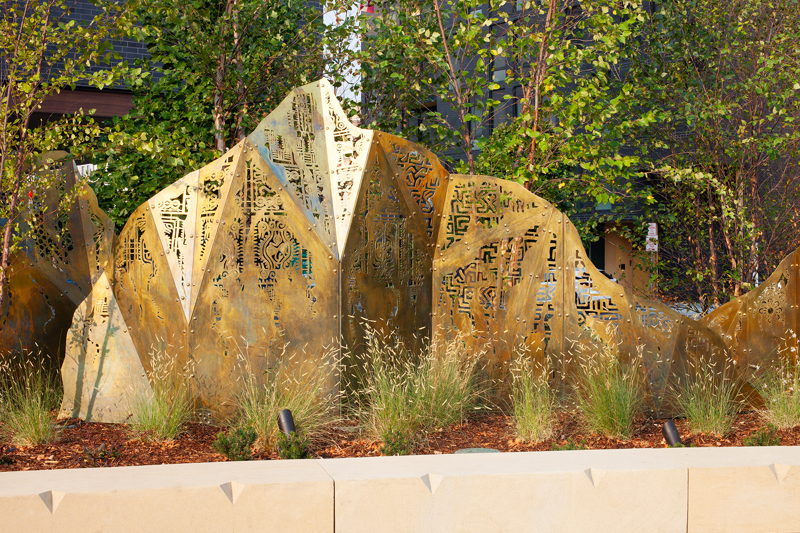 A geometric brass sculpture of mountain peaks topped with laser cut fabric patterns from various cultures. 