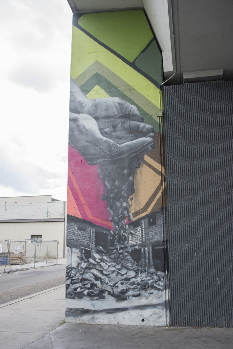 A mural painted in three parts on the wall of a highway overpass. On the left is the image of a pair of hands holding soil that slips through its fingers, turning into building rubble. In the middle is an image of a single eye situated beneath the arch of a bridge. On the right is an image of a hand and forearm turning into the crumbling facade of an abandoned building. 