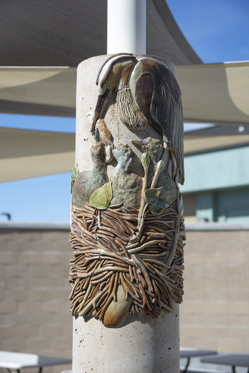 A cement column decorated with ceramic mosaic relief sculptures of a nesting great blue heron feeding chicks.