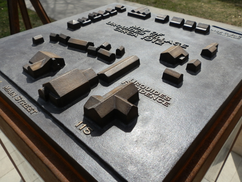 A life-size bronze sculpture of an antique tripod camera. In the viewing window of the camera is a portrait of a man (Jesus Urquides). In front of the camera is a stone pedestal for people to sit on as though having their picture taken. Next to the sculpture is a weathered steel pedestal topped with a model map of a community of houses (the Spanish Village). On the sides of the pedestal is signage describing the history of both Jesus Urquides and the history of the Spanish community in Boise. 