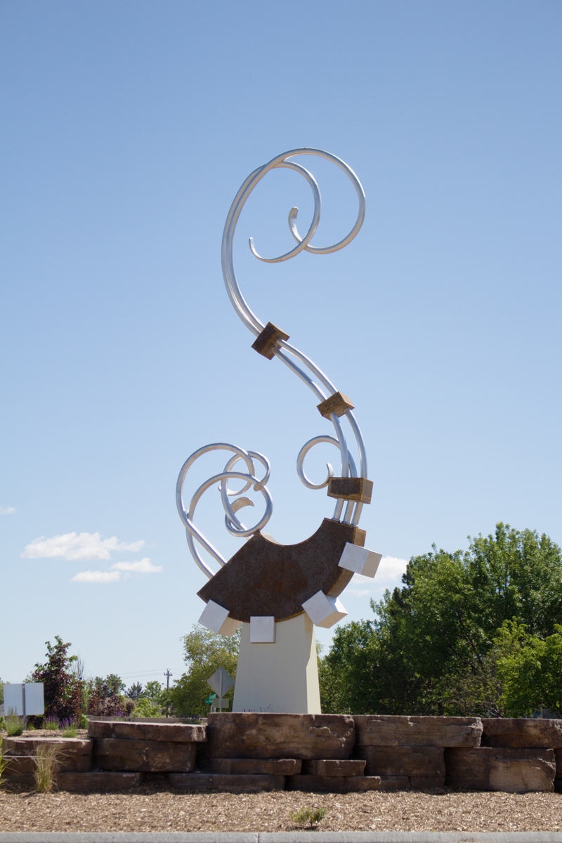 A twenty foot sculpture made of different metals including industrial steel, stainless steel, enameled steel, and aluminum. The sculpture is made of a number of aluminum pipes that curve and twist in the form of a letter S which intersect a number of square shapes along its center neck and base. 
