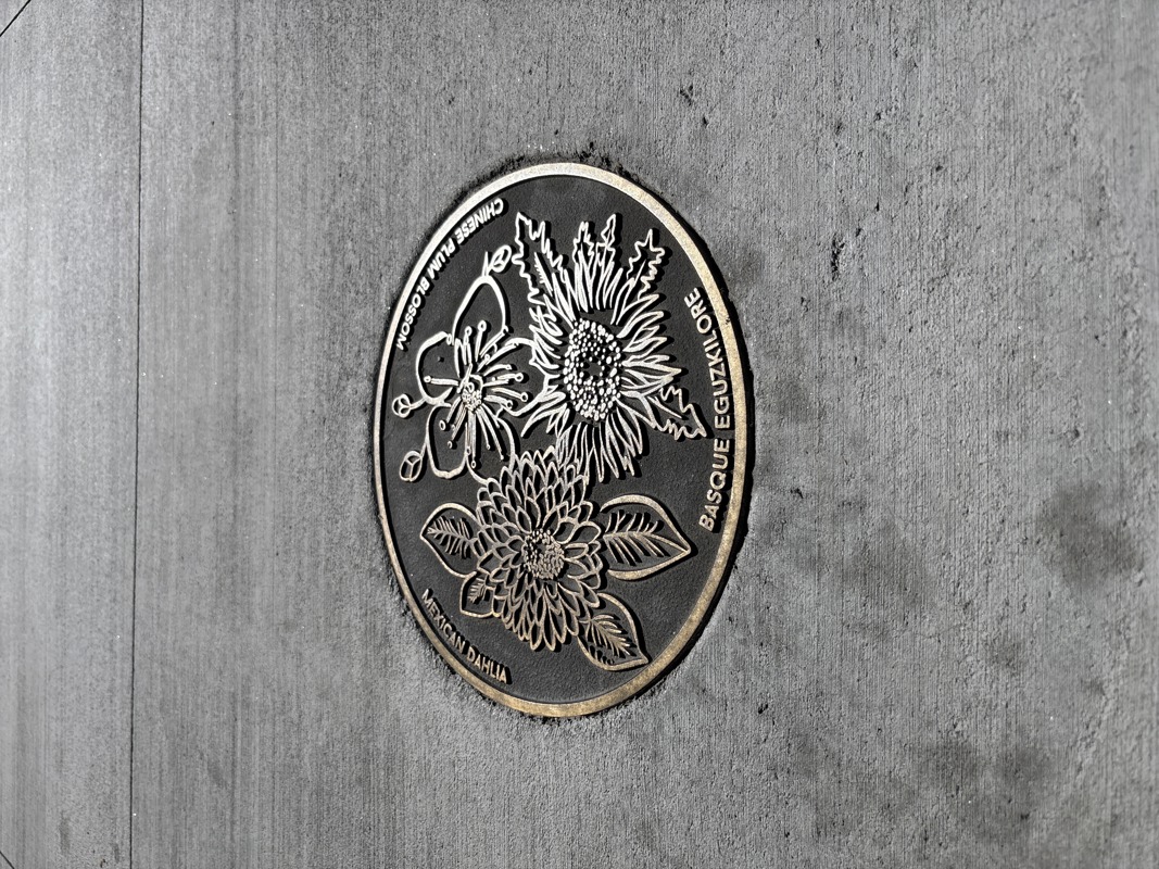 A bronze cast medallion inlaid in a cement sidewalk. The medallion features three flowers: a Basque eguzkilore, a Chinese plum blossom, and a Mexican dahlia.