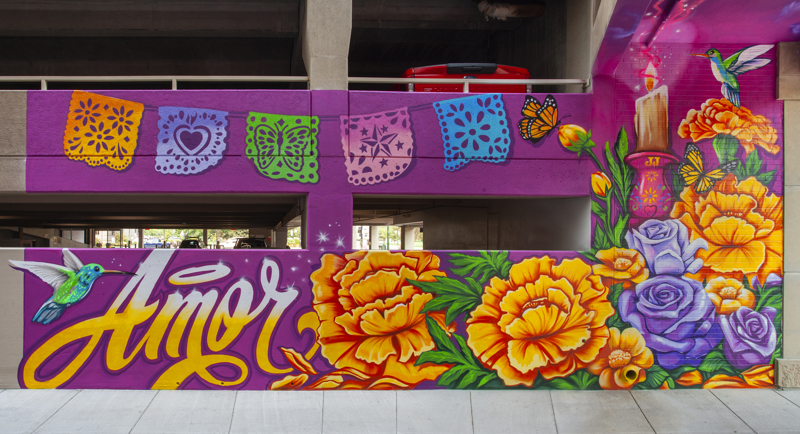 A painted mural that spans both sides of an alleyway. On one side is a dapperly dressed figure flying over the Boise skyline. They hold flowers and are surrounded by butterflies and swallows who carry notes of encouragement and affirmation. On the other side is an arrangement of flowers with butterflies, a hummingbird, a candle, and papel picado. On the far left is the word "Amore". 