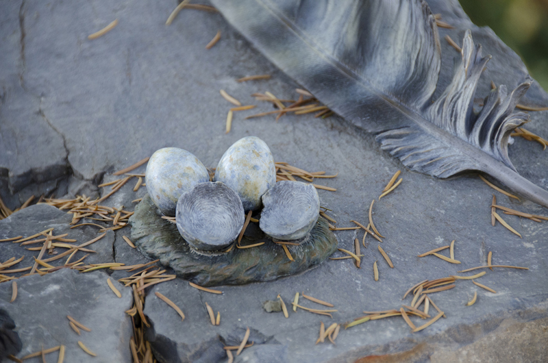 A detail closeup of a patinated bronze sculpture of a nest of eggs and a feather on top of a basalt stone surface.