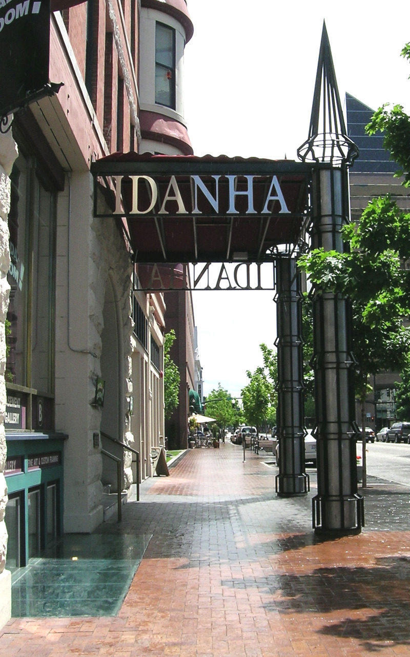 A building entrance canopy with the name "Idanha" at its top. The canopy is made of painted steel bars that create an impression of the Idanha Building's historical canopies. The canopies are held up by tower-like columns that mirror the building's existing turrets. Inside the columns are blue neon lights that illuminate them from within.  