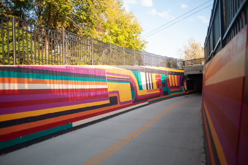 A mural on the retaining walls of a bike path heading into a tunnel. The design consists of lines and abstract shapes in the colors black, white, red, orange, yellow, teal, and magenta.