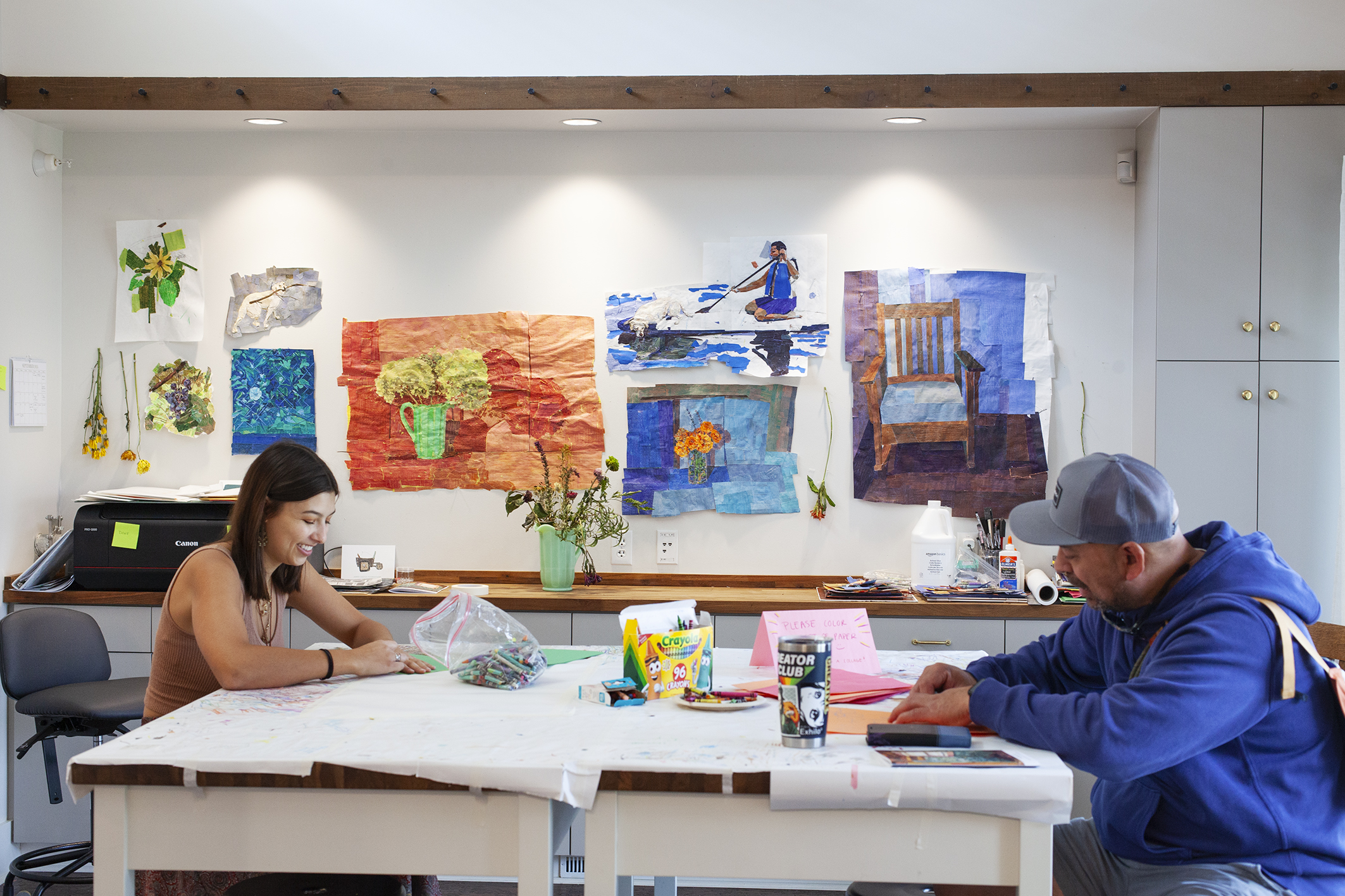 Murals hang on the wall behind an art table with two people creating art with crayons. 
