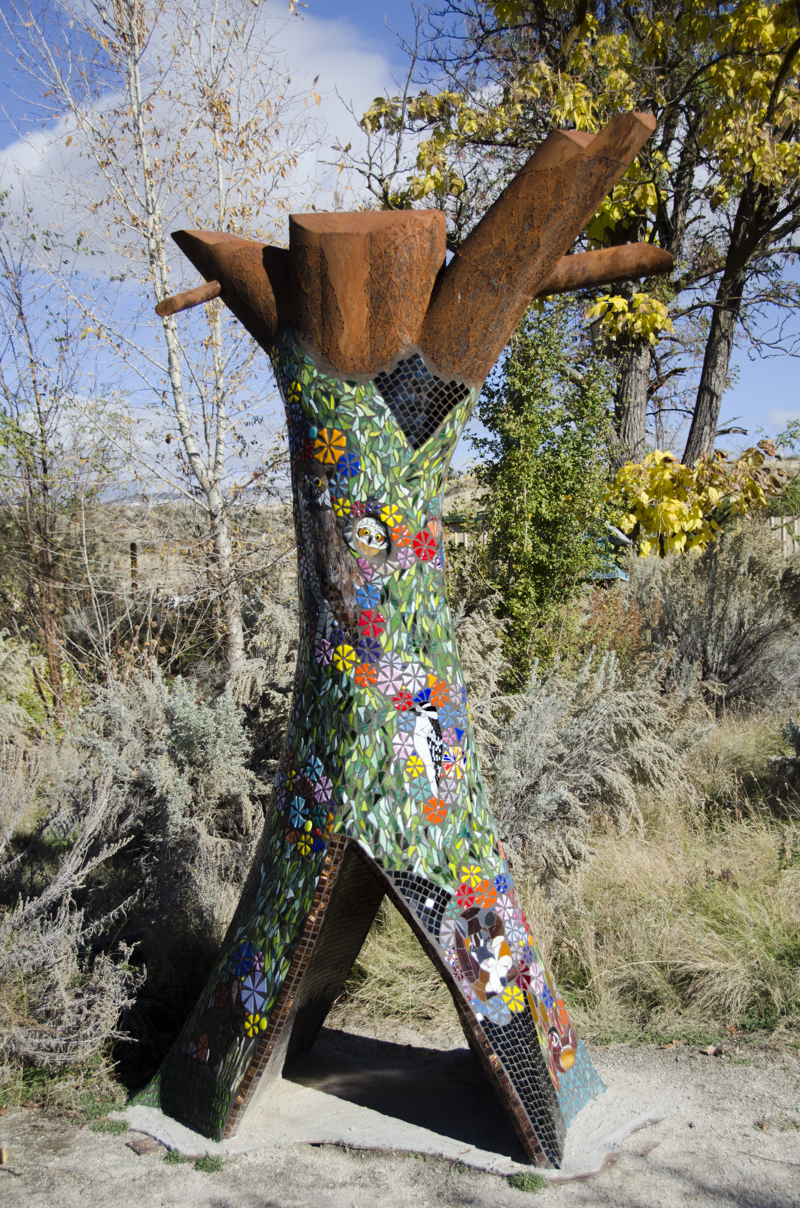 A life size sculpture of a dead standing hollow tree trunk. The sculpture is made of weathered steel and covered in glass tile mosaic depicting various lifeforms (i.e. birds, insects, and plants) that make use of dead or fallen trees in their habitat.