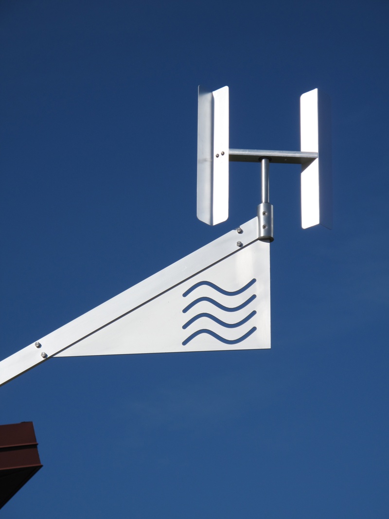 An aluminum kinetic sculpture representing a water molecule. At its central axis point is the letter O from which stems two arms with the letter H at their end. This images shows a detail closeup of the end of one of the arms which has a symbol of four wavy lines meant to symbolize the gaseous state of water.
