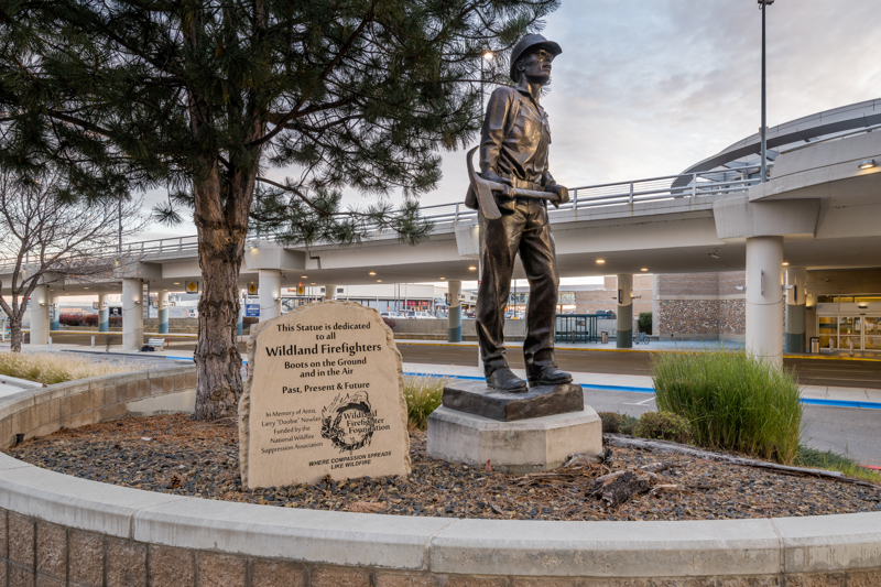 A bronze sculpture of a wildland firefighter wearing a helmet and holding a pulaski tool.