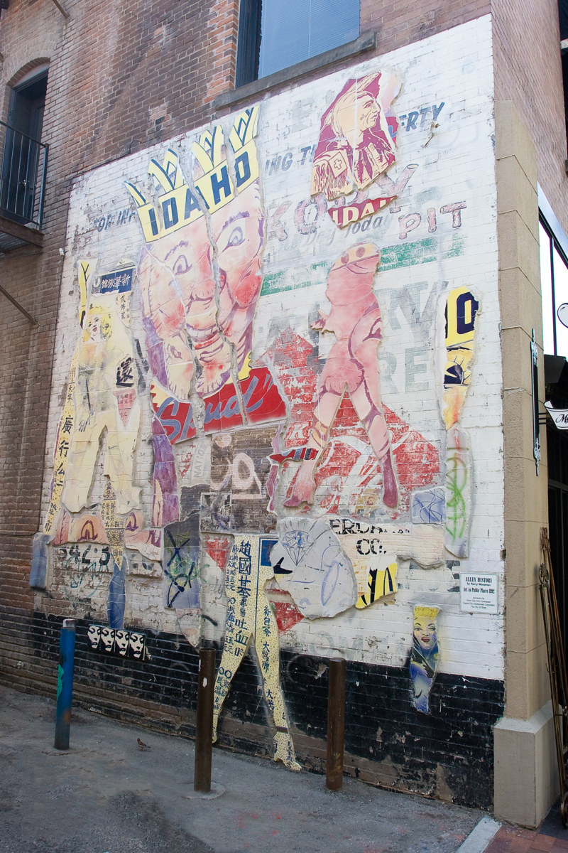 A mosaic made of fragmented glazed ceramic pieces installed on a brick wall at the corner of a building. On the ceramic pieces are a variety of symbols, images, and shapes referencing the location's history including Chinese characters, women, and old advertisements. Most prominently is a face wearing a crown with the accompanying text "Idaho" and  "Spud".