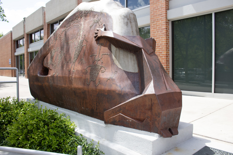 A weathered and stainless steel sculpture that depicts a Native American clinging to a rock formation that features petroglyphs. 