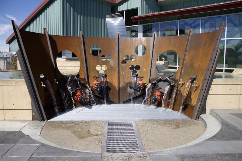 Fluxion is a sculpture that abuts a pool of water. The sculpture is made of 6 large curved plates of rusted industrial steel. In the plates are cut circular holes. Below the holes are several valve spickets of different sizes. Water from the pools spill through the holes and valves, and spills through a grate on the ground.