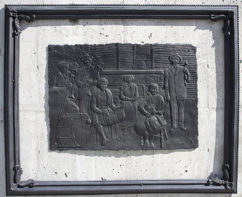 A low bas-relief bronze resin sculpture depicting a photo of 6 individuals with musical instrument. The sculpture is rectangular with a rough edge and is surrounded by a separate ornate frame.