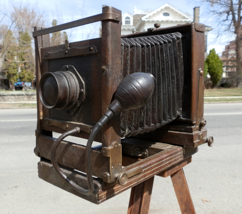 A life-size bronze sculpture of an antique tripod camera. In the viewing window of the camera is a portrait of a man (Jesus Urquides). In front of the camera is a stone pedestal for people to sit on as though having their picture taken. Next to the sculpture is a weathered steel pedestal topped with a model map of a community of houses (the Spanish Village). On the sides of the pedestal is signage describing the history of both Jesus Urquides and the history of the Spanish community in Boise. 