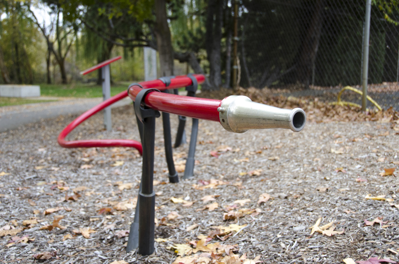 A long red pipe meant for sitting on. One end of the pipe coils out of the ground, while the other end has a fire hose nozzle. The pipe is held up by steel footings with clasps that resemble hands holding a firehose. 