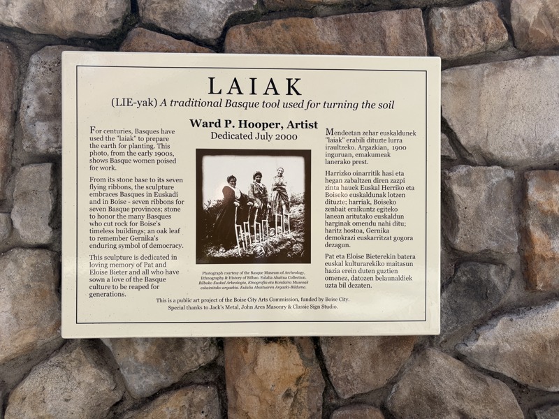 Signage which reads:

LAIAK
(LIE-yak) A traditional Basque tool used for turning the soil
Ward P. Hooper, Artist
Dedicated July 2000

For centuries, Basques have used the "laiak" to prepare the earth for planting. This photo, from the early 1900s, shows Basque women poised for work. 

From its stone base to its seven flying ribbons, the sculpture embraces Basques in Euskadi and in Boise - seven ribbons for seven Basque provinces; stone to honor the many Basques who cut rock for Boise's timeless 