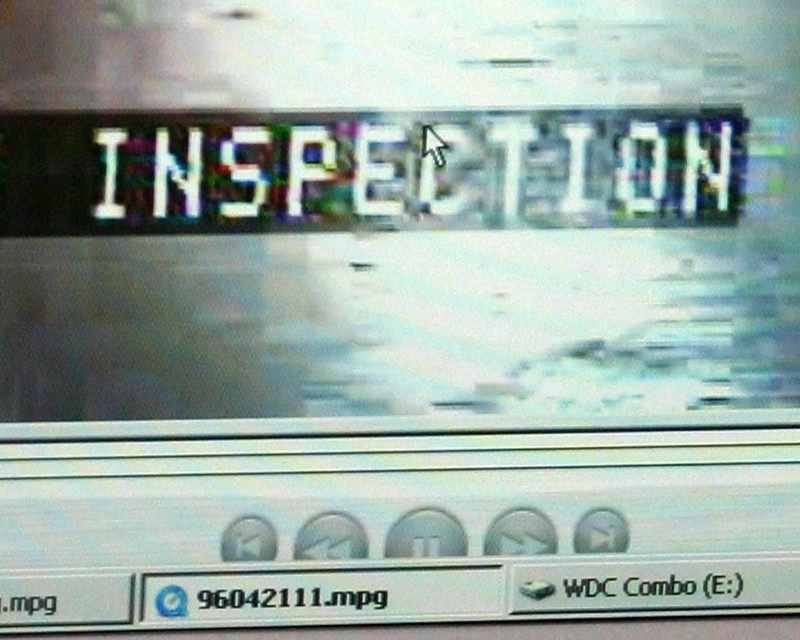 A closeup of a computer screen with a video window. In it is a black and white text ribbon with the word "INSPECTION". Over the words hovers a white arrow cursor. At the bottom of the video window are the play option buttons. Below them is the computer screen task bar.  