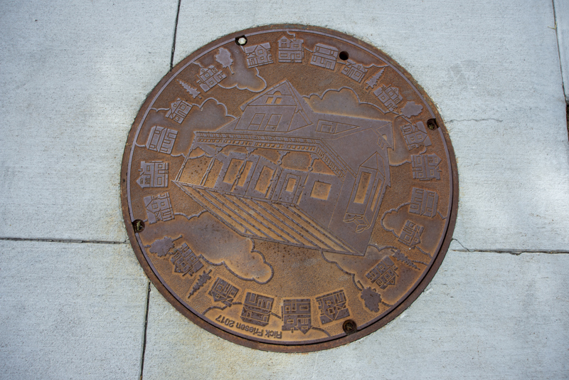 An iron utility (or man hole) cover with a relief sculpture of a floating Victorian house. Along the edge of the cover is a boarder of houses and trees.