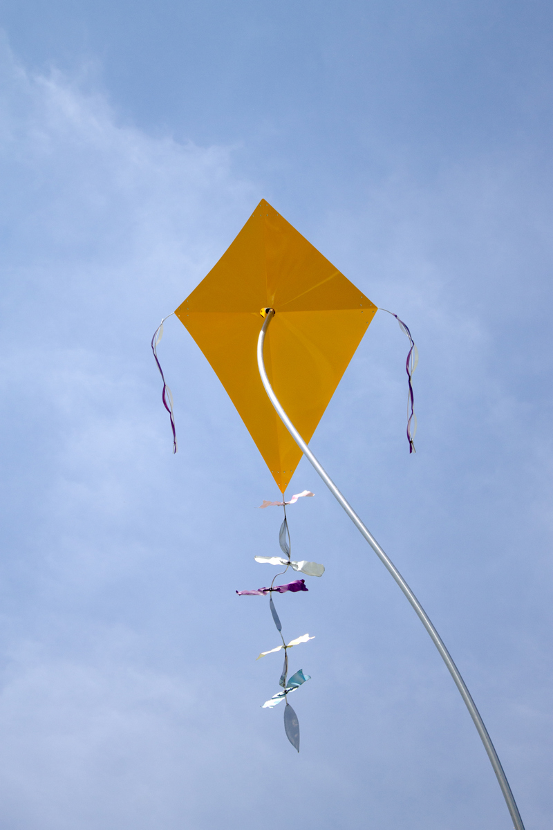 A closeup of one of the kites which is yellow and in the traditional diamond shape with bows tied along its tail.