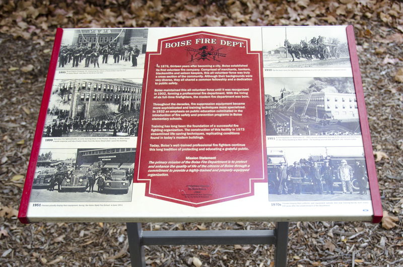 A long red pipe meant for sitting on. One end of the pipe coils out of the ground, while the other end has a fire hose nozzle. The pipe is held up by steel footings with clasps that resemble hands holding a firehose. 