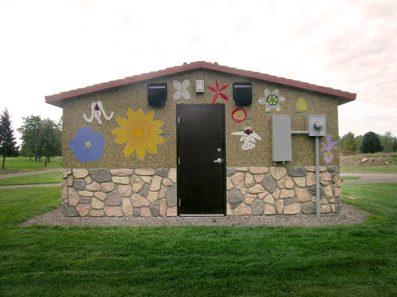 A small park bathroom building with sides covered in tile mosaics depicting flowers found in the state of Idaho. 