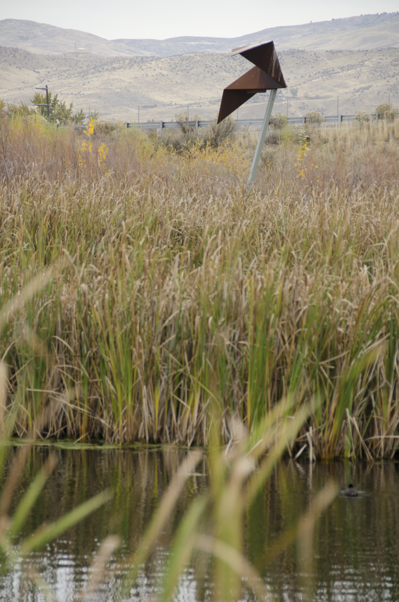 An abstracted geometric sculpture resembling a paper-folded origami bat in flight made of weathered steel, mounted onto a pole in the middle of a wetland.