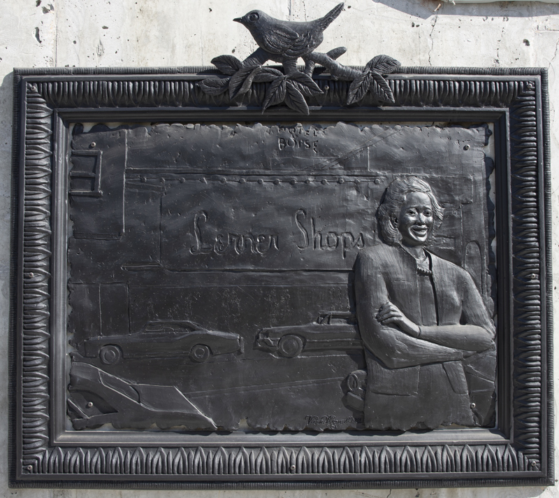 A low bas-relief bronze resin sculpture depicting a woman standing in front of a business sign which reads, "Lerner's Shops". The sculpture is rectangular with a rough edge and is surrounded by a separate ornate frame which has a bird on its top center.