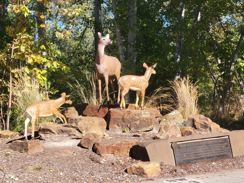 A painted sculpture of a deer and two fawns standing on a hill of rocks. Beneath them is a bronze plaque.