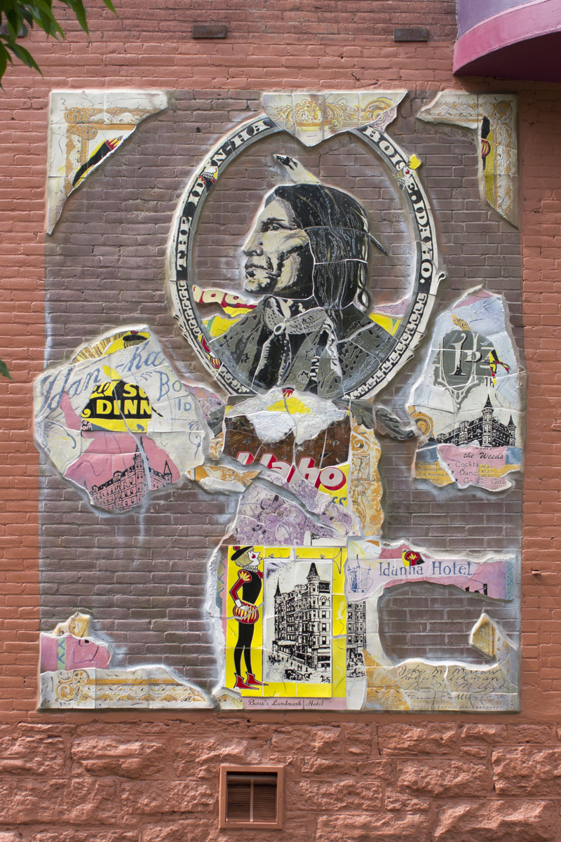 A mosaic made of fragmented glazed ceramic pieces installed on a brick wall painted red. On the ceramic pieces are images of the historic Idanha building. Featured at the top center is a profile portrait of a Native American.
