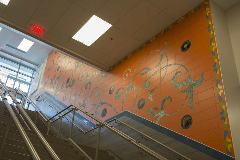 A mosaic wall installation made of ceramic tile. Made with green glass tile are leafy vines. Interspersed are tiles with printed images of local native flowers.