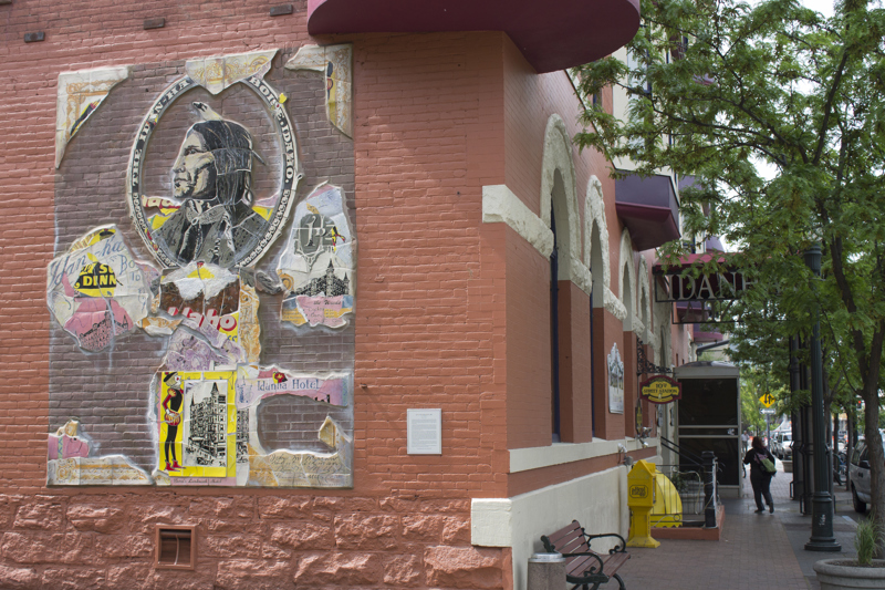 A mosaic made of fragmented glazed ceramic pieces installed on a brick wall painted red. On the ceramic pieces are images of the historic Idanha building. Featured at the top center is a profile portrait of a Native American. 