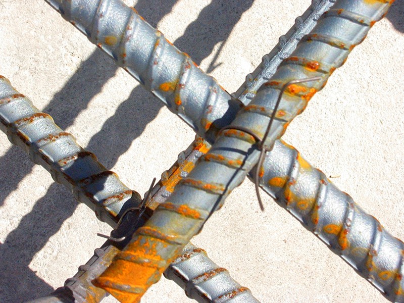 A closeup photograph of four intersecting pieces silver-painted rebar tied together with wire. A vibrant orange corrosion has formed on the rebar.