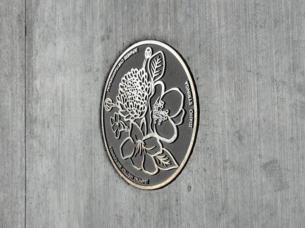 A bronze cast medallion inlaid in a cement sidewalk. The medallion features three flowers: an Idaho syringa, a Japanese chrysanthemum, and a Congolese Yellow Trumpet. 