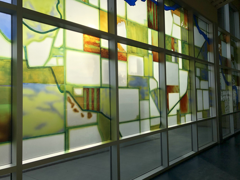 A window installation resembling stained glass depicting a birds-eye view of a blue river flowing through the geometric rectangular shapes, some that looks like fields and housing communities.