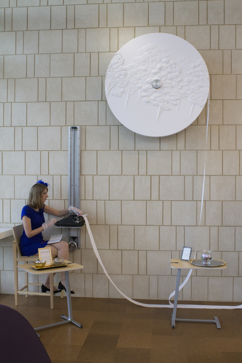 An antique typewriter set atop of a shelf that can be raised or lowered along a crankshaft attached to a sandstone wall. The typewriter is fed from a large roll of paper also attached to the wall. The surface of the paper roll features a low-bas relief carving of poplar trees. The typewriter has been lowered and a person in a blue dress is using it to type.