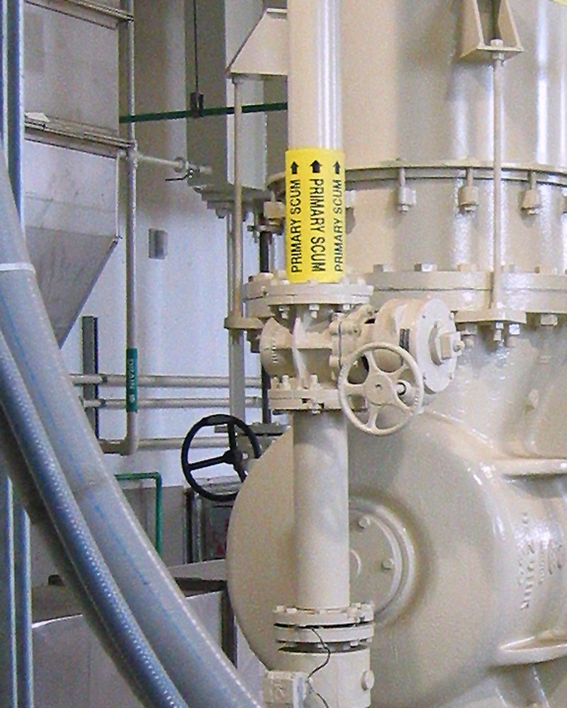 Heavy indoor water treatment machinery painted white. There are a number of large tubes, pipes, and valves. On one pipe is a yellow label with the text, "PRIMARY SCUM," with an upward pointing arrow.