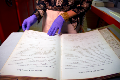 Gloved hands begin to turn the page on a large cemetery deed book with large scripted letters.