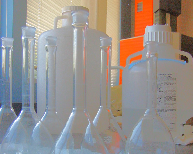 A closeup of six empty long-neck volumetric flasks used in chemistry. Behind them are two large white plastic chemical jugs.