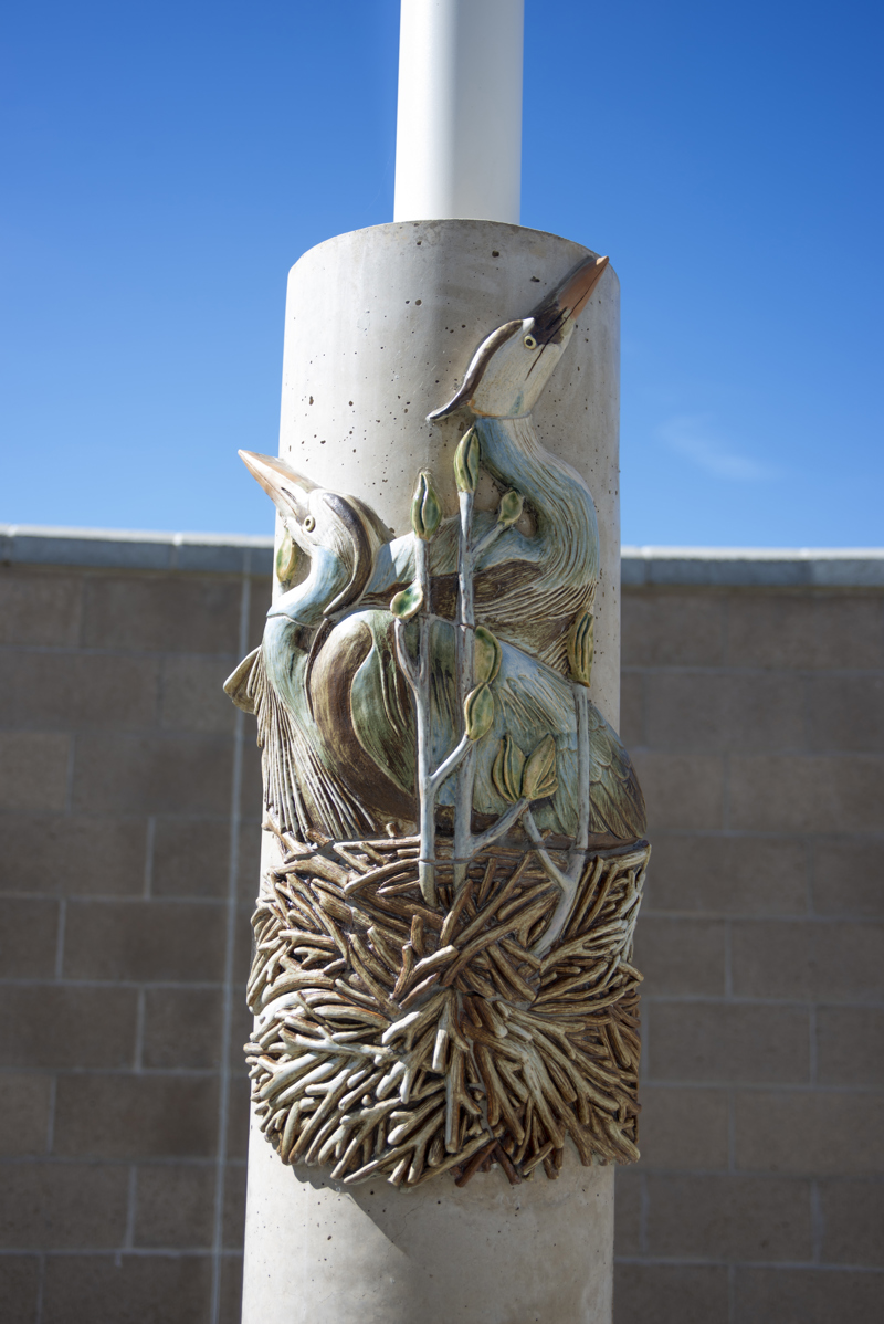 A cement column decorated with ceramic mosaic relief sculpture of a nesting pair of great blue herons.