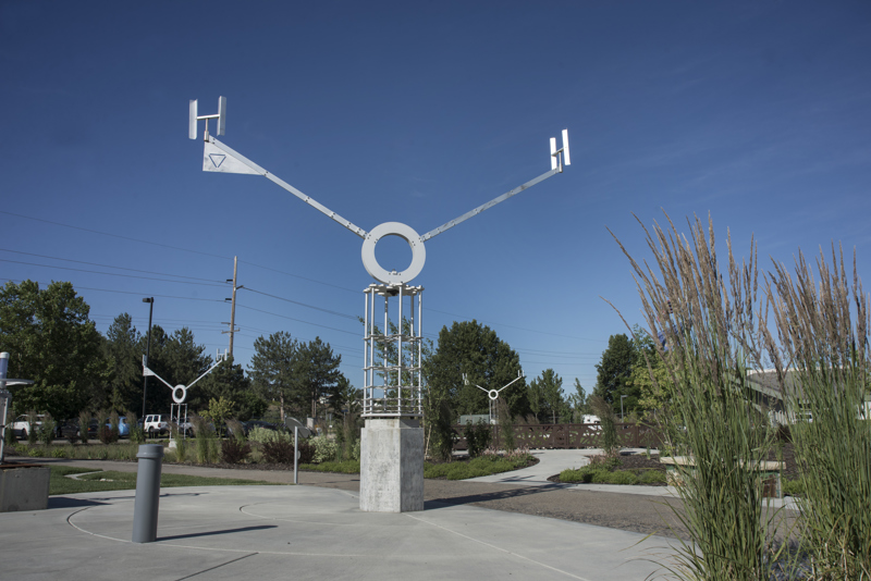 An aluminum kinetic sculpture representing a water molecule. At its central axis point is the letter O from which stems two arms with the letter H at their end. This sculpture turns in the wind like a weather vane and sits atop a column made from a stack of aluminum plates cut in the shape of snowflakes. 