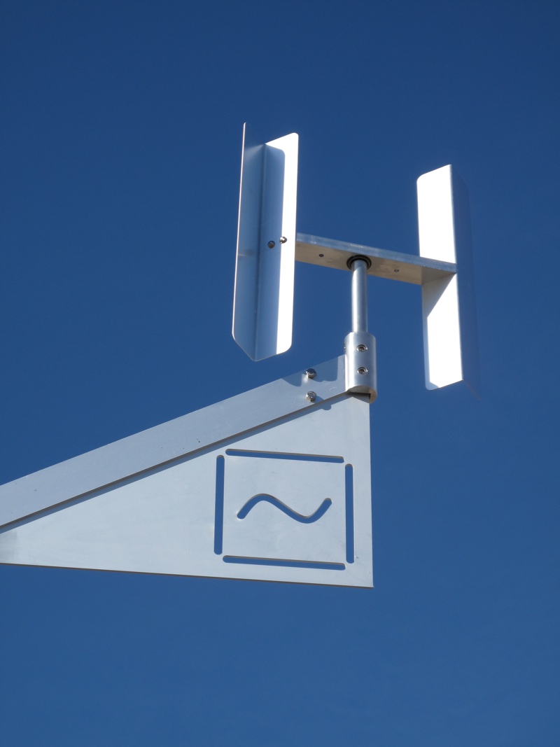 An aluminum kinetic sculpture representing a water molecule. At its central axis point is the letter O from which stems two arms with the letter H at their end. This images shows a detail closeup of the end of one of the arms which has a symbol of a single wavy line in a square meant to symbolize the liquid state of water.