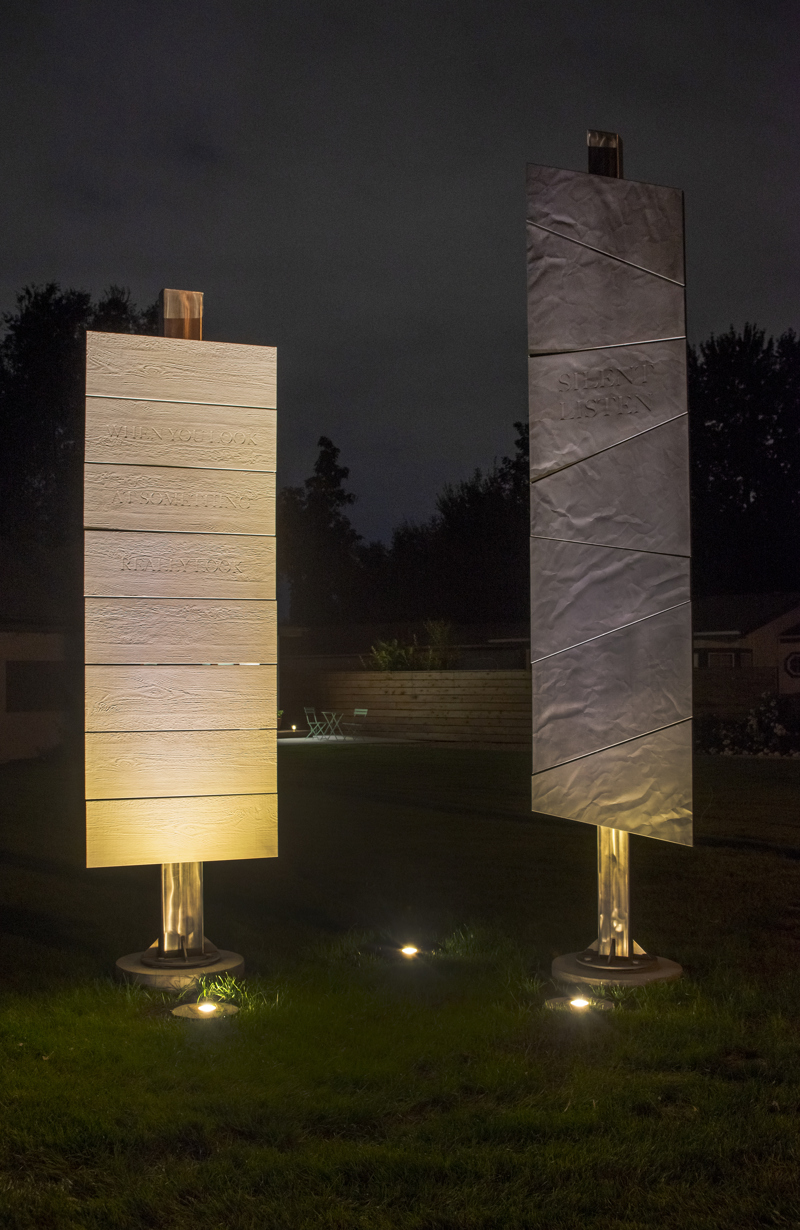 Two standing sculptures made of cement panels affixed to steel poles. One sculpture appears white with the cement panels looking like 8 horizontal planks of wood with sunken text that reads, "When you look at something, really look." The other sculpture appears black with slanted cement panels that have the surface texture of flagstone with sunken text that reads, "Silent listen." 
