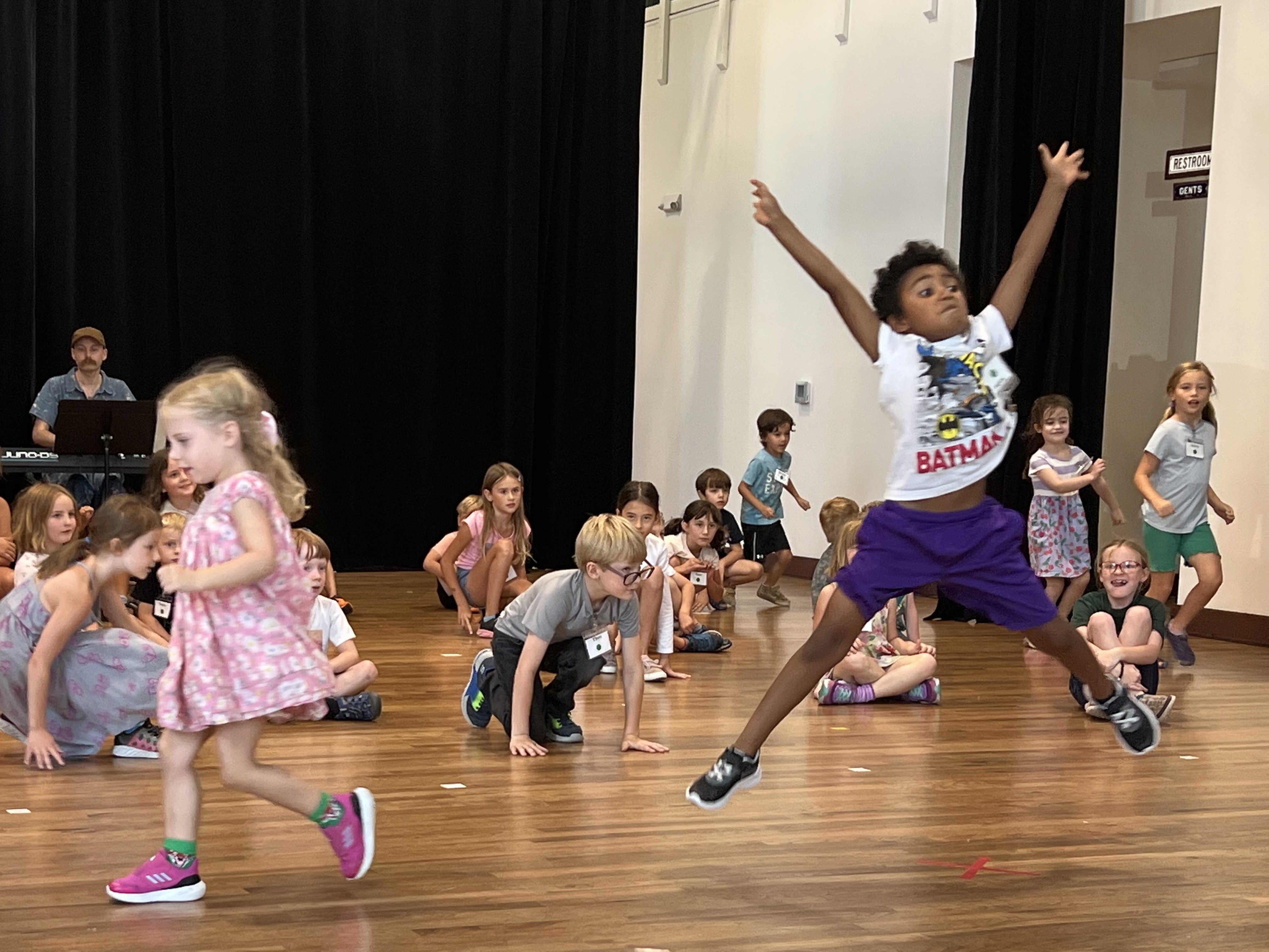 Kids in line dancing, one leaping through the air, piano accompanist providing the music. 
