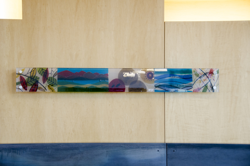 A small rectangular installation of colorful fired glass mounted onto the wall above a fireplace. On the glass are images of animals and plants referencing the area's agricultural history.