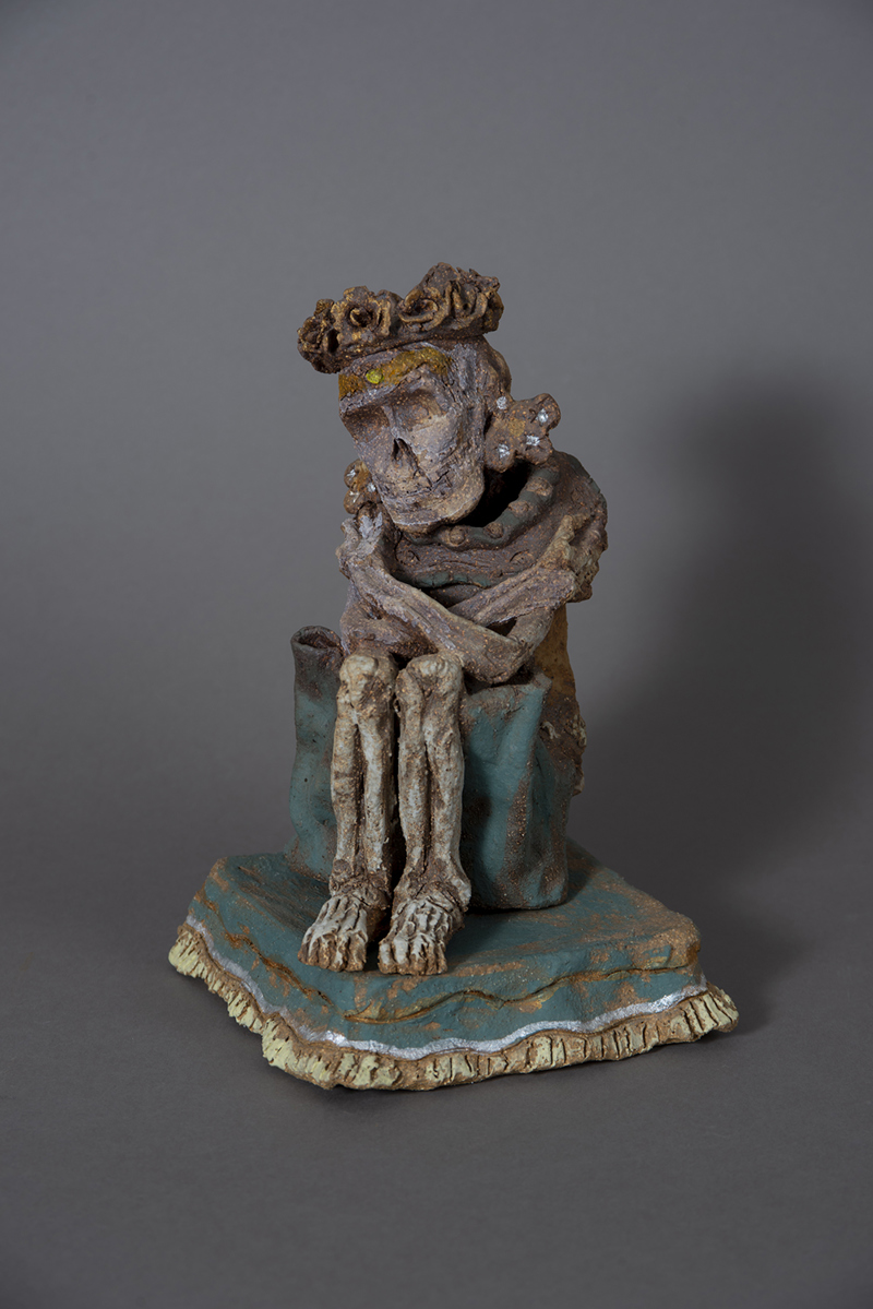 A sculpture of a skeleton with crossed arms seated on a pillow with fringe. On the skeleton's head is a crown of roses.