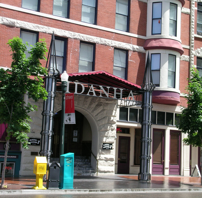 A building entrance canopy with the name "Idanha" at its top. The canopy is made of painted steel bars that create an impression of the Idanha Building's historical canopies. The canopies are held up by tower-like columns that mirror the building's existing turrets. Inside the columns are blue neon lights that illuminate them from within.  